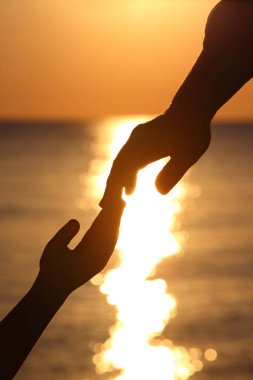 Gün batımında deniz kenarında iki elin silueti, çocuk ve anne parmaklarını birleştirir.