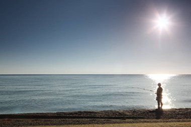 Balıkçı silueti kıyıda duruyor ve balık avlıyor.