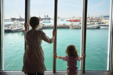 woman and her daughter standing and looking at ships in Qaboos Port through glass. clipart