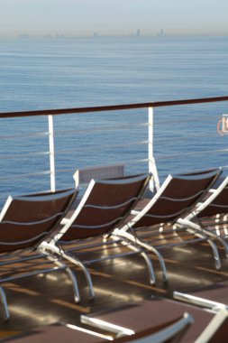 Yolcu gemisinin güvertesindeki bir çok şezlong odak dışı. Uzaktaki binalar.