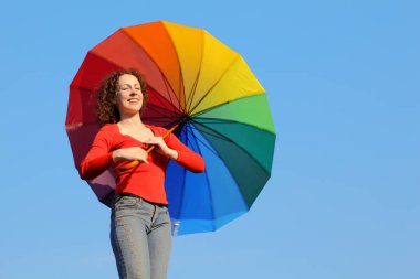 Güneş parladığı için gözleri kapalı bir kız mavi gökyüzüne karşı duruyor ve elinde renkli bir şemsiye tutuyor.