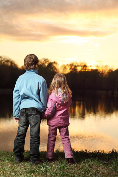 Little girl in pink clothes and boy in blue jacket standing back on bank of river and admire on sunset