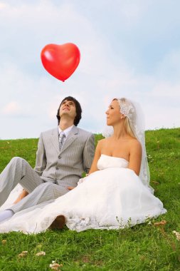 Güzel genç damat ve gelin beyaz elbiseler giyip yeşil çimlerde oturuyor ve kalp şeklinde kırmızı balon uçuşunu izliyorlar.