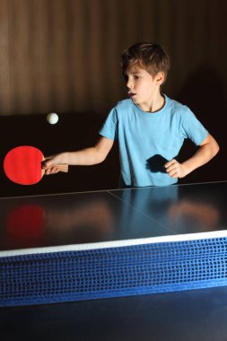 Mavi tişörtlü küçük çocuk ping pong oynuyor; konsantre yüz; mavi ağ