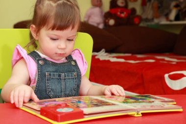 Kot tulumlu, pembe tişörtlü, sarı koltuklu güzel küçük kız kitap okuyor.