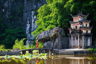 Beautiful woman wearing red dress and conical hat visiting Bich Dong pagoda in Ninh Binh Vietnam. Bich Dong Pagoda is a popular tourist destination of Asia. clipart