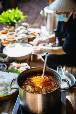 A pot of simmering broth with meat and vegetables, part of a Bun Cha preparation. Nearby, a cook arranges fresh ingredients and noodles in a rustic kitchen setting clipart