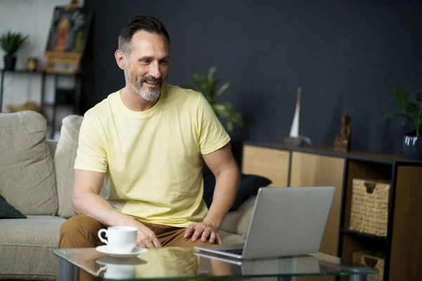 ソファーに座っている中年男性とオンライン会議を持っているラップトップを使用して ソファコーヒーを飲みながらガラステーブルの横にあるソファ 成熟したハンサムなフリーランスItマン仕事上のコンピュータ自宅 — ストック写真