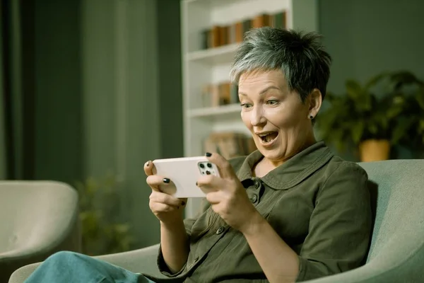 Woman at home is fully engrossed in her phone game, with a big smile on her face as she plays. Her happiness is evident as she focuses on achieving her goals in the game. High quality photo