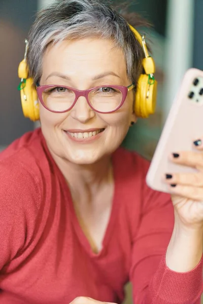 Mature woman wearing glasses, smiling and holding phone headphones on. Image portrays technology, communication, and entertainment, while womans happy expression reflects her enjoyment and