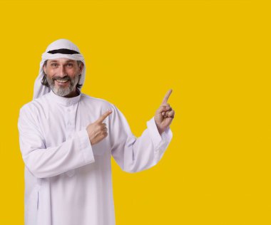 Smiling Arab man captures his cheerful and confident personalit, he points both hands towards copy space on isolated yellow background. Copy space for designers and publishers to add their text, logos clipart