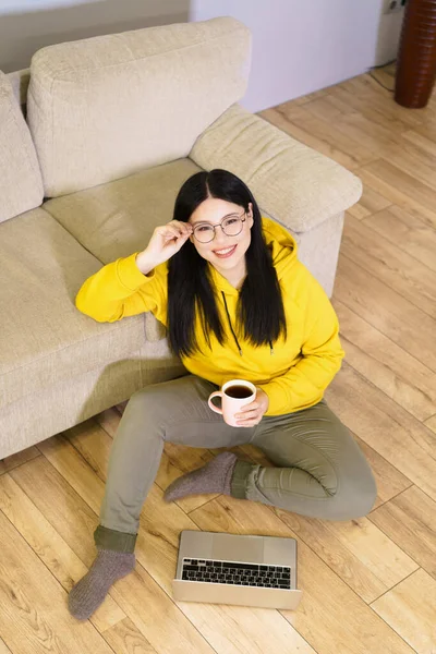眼鏡をかけた若いアジア人女性が 目の前にノートパソコンを持ち コーヒーを飲みながら床に腰を下ろしている 彼女はデジタル作品に従事しており リラックスした瞬間を楽しんでいます — ストック写真