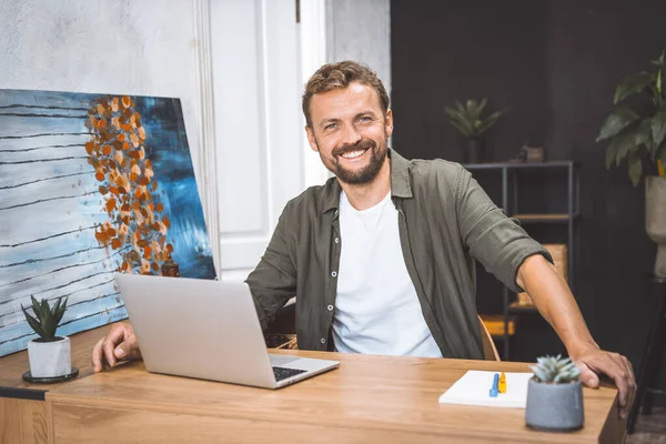 Concept of remote work, freelance, and productivity in digital age. Cheerful and productive man, works from home, He smiling, focusing on his work, surrounded by digital technology and equipment
