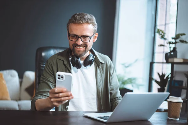 幸せと笑顔の男はロフトオフィスに座っている カジュアルな服装で身を包んだ 彼の携帯電話にメッセージをテキストメッセージしながら 彼のラップトップで作業 高品質の写真 — ストック写真