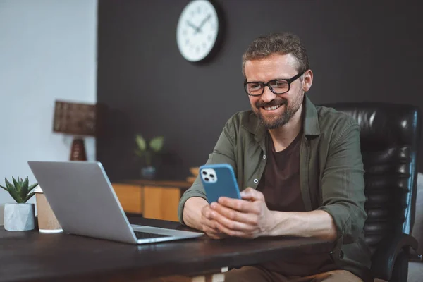 笑顔と幸せな中年の男性はノートパソコンの近くの机の上に家に座っている間 オンライン活動に従事しています 男性は テキストメッセージを送るか 電話でオンラインで時間を過ごすかのどちらかで マルチタスクを描写されます — ストック写真