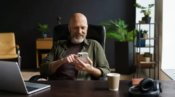 灰色のひげを生やした大胆かつ立派な男 彼は喜びの瞬間を捉え 電話でメッセージを読みながら笑顔を浮かべた テーブルにはコンピュータ コーヒーカップ オーディオヘッドフォンがあります 高品質 — ストック写真
