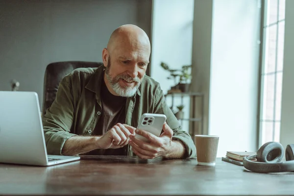 熟女老人は快適に自宅に座って 積極的に電話でテキストメッセージに従事した ノートパソコン ヘッドフォン コーヒーカップで装飾されたデスクは 仕事やリラクゼーションに役立つセットアップを示しています そうだ — ストック写真