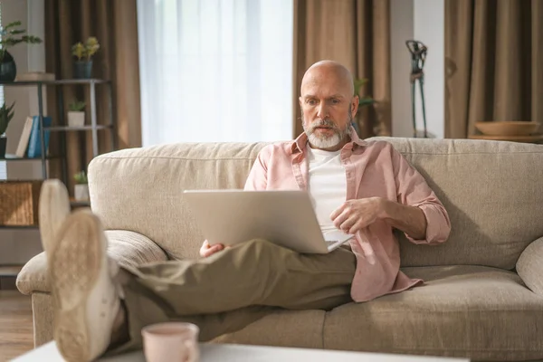 時間を楽しむ姿を描いた銀髭の老人 彼は快適にソファに座り ノートパソコンでインターネットを見ることに夢中になった 高品質の写真 — ストック写真
