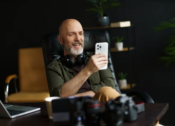 中年男人 银胡子 沉迷于在手机上看新闻 将笔记本电脑放在书桌上作为背景图 图像捕捉了连通和数字化生活方式的本质 — 图库照片