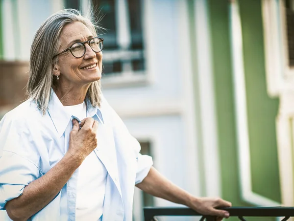 老年快乐 成熟的女人带着灿烂的笑容站在阳台上俯瞰着欧洲的老街 老太太心情愉快 心满意足 优雅地拥抱着老年之美 — 图库照片