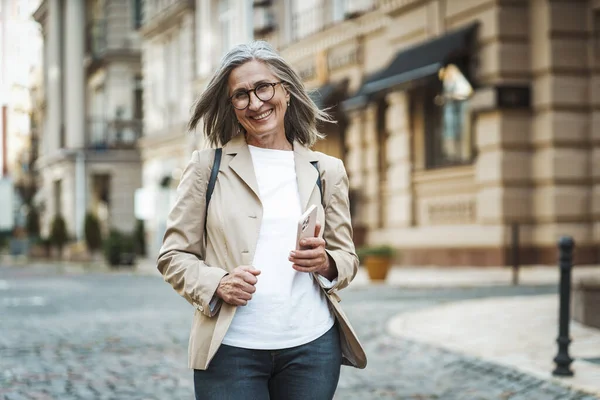 一位快乐的老年女士在欧洲老城区的街上被抓获 手里拿着纸杯里的咖啡 笑容可亲的成熟女士 一头银发 优雅地走在迷人的街道上 是的高质量的照片 — 图库照片