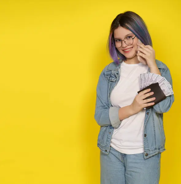 Financial Independence Associated With Managing Money In Modern And Confident Manner. Happy Smiling Teenager Girl With Wallet Full Of Money And Mobile Phone Isolated On Yellow With Copy Space. High