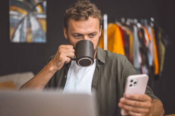 Ein Mann Blickt Auf Sein Handy Während Einen Kaffeebecher Hält lizenzfreie Stockbilder