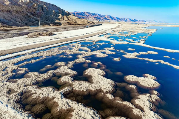 The blue water is surrounded by pink mountains. The picturesque Dead Sea is covered with patterns of salt. The picture was taken by a drone from a aerial view. Israel. Winter sunny day.