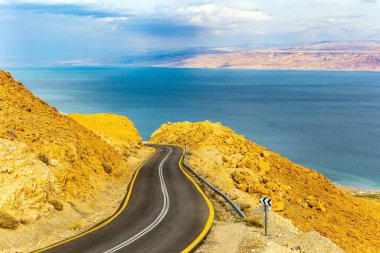 Otoyol Ölü Deniz kıyısı boyunca uzanıyor. İsrail. Denizin karşı kıyısındaki Ürdün loş kış güneşiyle aydınlanır. Bulutlu Ocak günü. Deniz yüzeyi bulutları yansıtır..