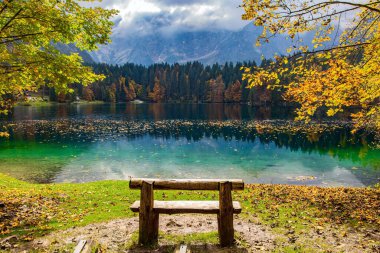 Kuzey İtalya 'da buzul kökenli Lago Fusine. Göl kenarında ahşap bir bank. Suyun pürüzsüz yüzeyi ormanları yansıtır. Dağlar sabah kaybıyla kaplandı
