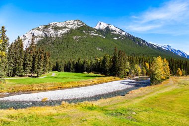 Kanada 'nın Rocky Dağları. Rundle ve Cascade Dağları 'nın eteklerindeki golf sahaları. Dağlar yoğun çam ormanlarıyla kaplı. Kıçı çakıllı, sığ bir dere.. 