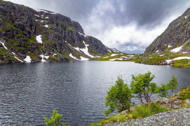 Büyük kar tarlaları gölün suyuna yansıyor. Norveç 'te soğuk bir yaz. 520 nolu yolda karavanla seyahat ediyorum. Geçen yılın karı ve gri gökyüzü. Şiddetli gri bulutlar gökyüzünü kaplıyor. 