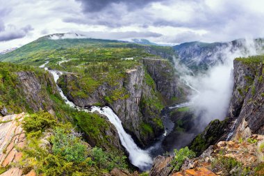 Norveç 'te soğuk Temmuz yağmurlu. Voringsfossen, Hardangerfjord 'daki görkemli şelaledir. Balık gözü merceğiyle çekilmiş bir fotoğraf. Şelale dar ve derin bir vadiye düşüyor..