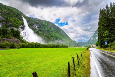 Roaring high-water cascading waterfall Vidfossen. Lush clouds and constant cold rain. Scenic road in the mountains of Western Norway. White water foam envelops the foot of the waterfal