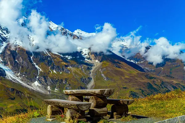 Snowy mountain peaks covered with clouds. The famous mountain road Grossglognerstrasse. Wooden table and picnic benches. Austria. The Hohe Tauern Park.