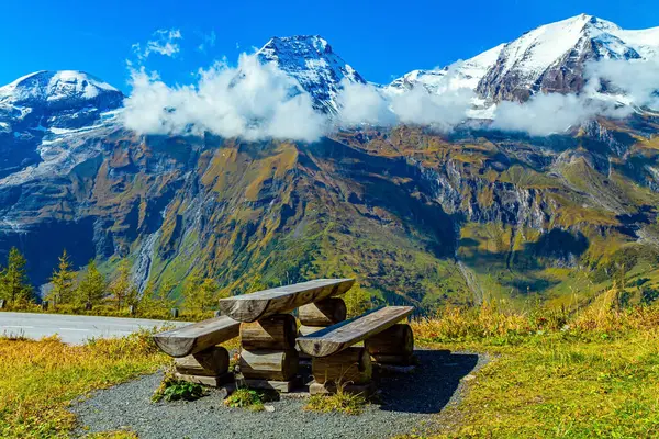 Wooden table and picnic benches. The famous mountain road Grossglognerstrasse. Austria. The Hohe Tauern Park. Snowy mountain peaks covered with clouds.