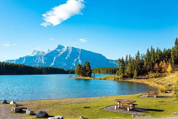 Cozy wooden picnic table and benches. Two Jack Lake. Huge glacial lake reflects the sun and clouds. Travel to Canada, the Rocky Mountains.