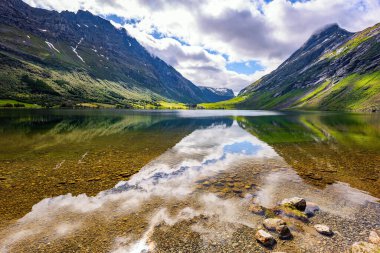 Temiz şeffaf suyu olan sığ bir göl. İskandinavya 'da yaz mevsimi. Eidsvatnet - Norveç 'teki görkemli göl. Su yemyeşil bulutları, mavi gökyüzünü ve ormanlı kıyıları yansıtır.. 