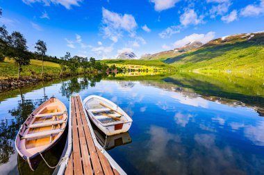 Wooden pier with boats. Shallow lake Eidsvatnet in the mountains of Norway. Summer in Scandinavia. The water reflects lush clouds, blue skies and forested shores.  clipart