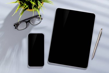 Ofis masasında boş ekranlı tablet ve akıllı telefon. Güneşli ışıklandırmalı düz çalışma alanı ve kopyalama alanı