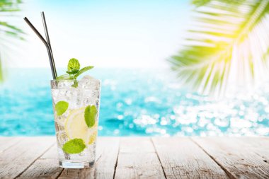 Güneşli deniz ve palmiye yapraklarının önündeki ahşap masada mojito kokteyli.