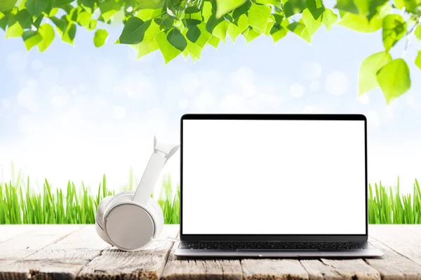 Laptop with blank screen and headphones on wooden table in front of sunny landscape. Work and travel or remote business concept