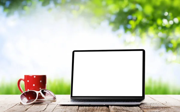 Laptop with blank screen and coffee on outdoor wooden table in front of sunny bokeh. Outside location education and business concept