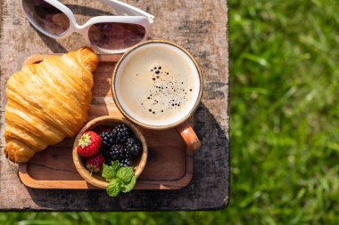 Kahve fincanı ve kruvasan bahçe masasının üstünde. Güneşli bir açık hava yemeği. Düz yatıyordu