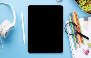 Boş ekranlı tablet, okul malzemeleri, kırtasiye malzemeleri ve mavi arka planda beslenme çantası. Eğitim ve beslenme. Metnin ya da uygulaman için boş alanı olan düz uzan