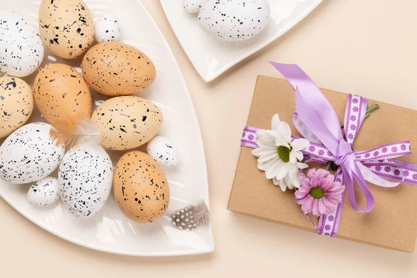 Easter eggs and flowers on plate and gift box over beige background. Flat lay
