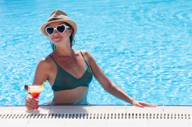Kalp şeklinde güneş gözlüklü bir kadın tropik bir kokteyl eşliğinde havuzda dinleniyor. Kopyalama alanı ile