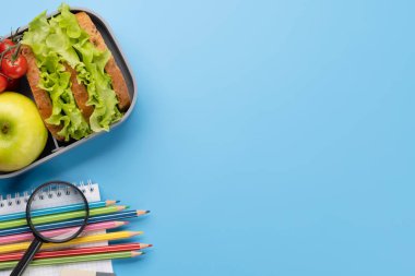 School supplies, stationery, and lunch box on blue background. Education and nutrition. Flat lay with blank space