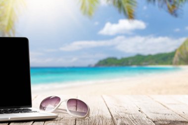 Dizüstü bilgisayar ve güneş gözlükleri güneşli denizin ve palmiye yapraklarının önündeki ahşap masada. İş ve seyahat ya da uzak iş konsepti