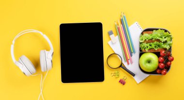 Boş ekranlı tablet, okul malzemeleri, kırtasiye malzemeleri ve sarı arka planda beslenme çantası. Eğitim ve beslenme. Metnin ya da uygulaman için boş alanı olan düz uzan
